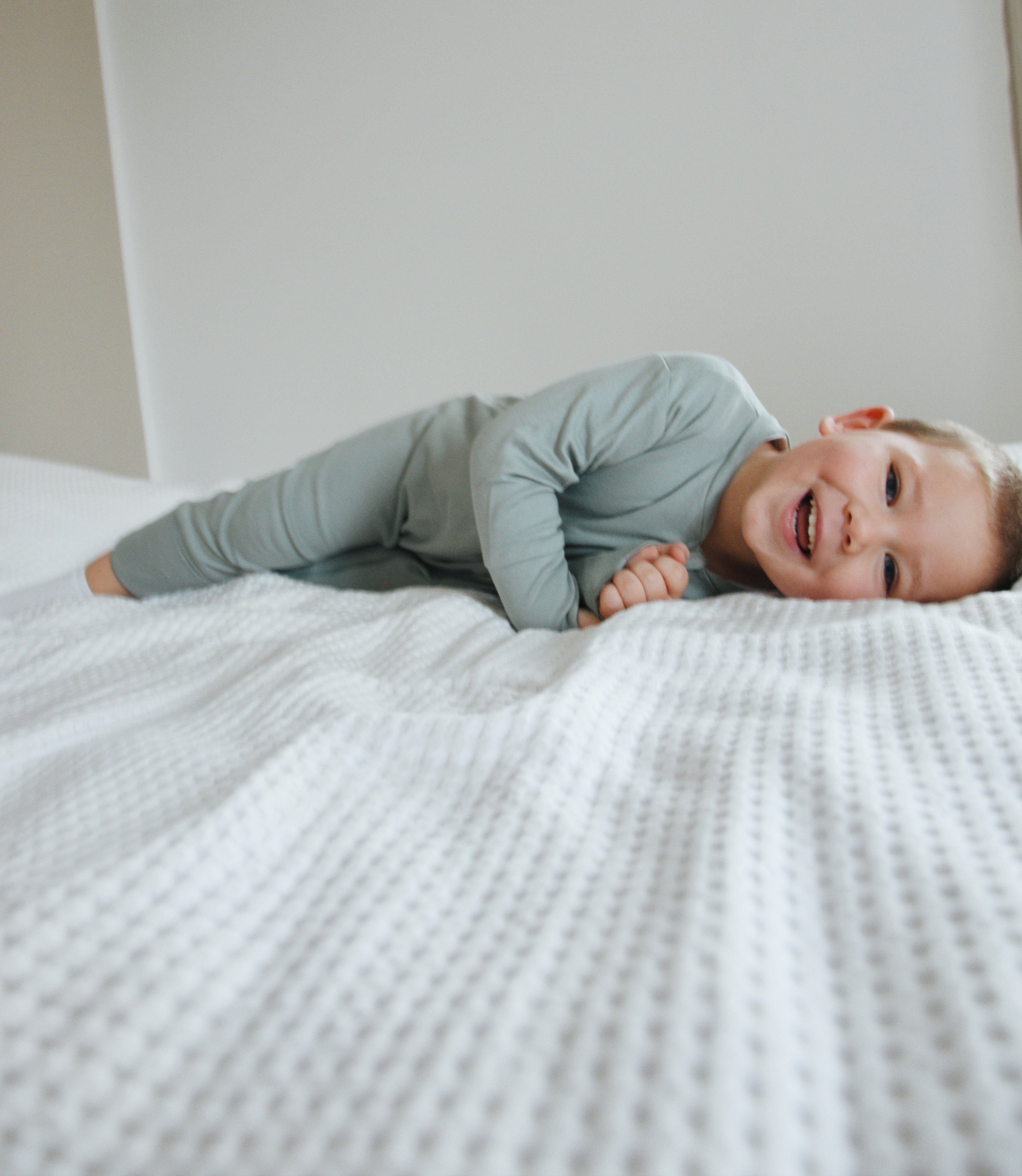 Lachendes Kleinkind liegt auf weißem Bett mit Bio-Babymode in gedecktem Grün von Elsara