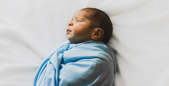 Neugeborenes Baby in hellblauer, biologischer Babymode liegend, sanft eingewickelt auf weißem Untergrund