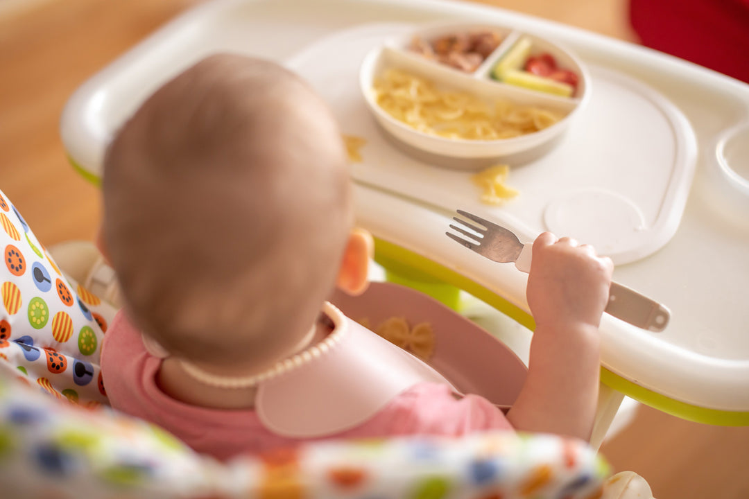 Einjährige Baby in Hochstuhl mit pinkem Lätzchen hält Gabel, vor Teller mit Pasta und Gemüse