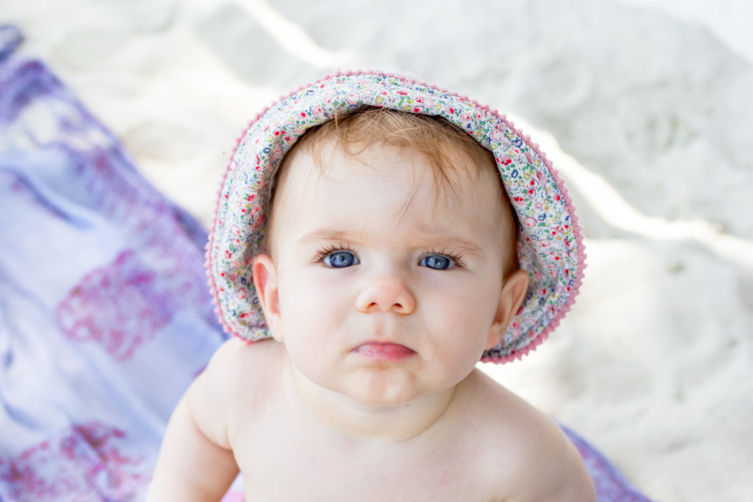Nahaufnahme eines Babys mit blauen Augen und Blumensonnenhut am Strand auf einem Tuch
