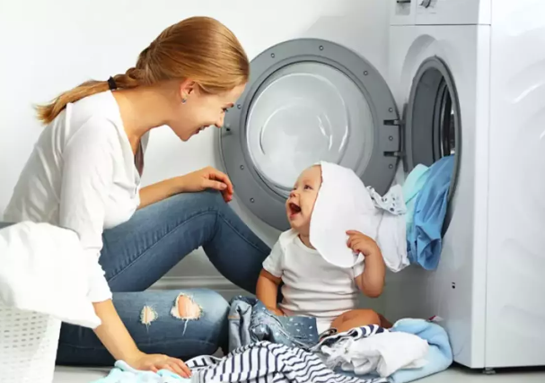 Mutter spielt mit lachendem Baby vor Waschmaschine mit Bio-Babykleidung von Elsara