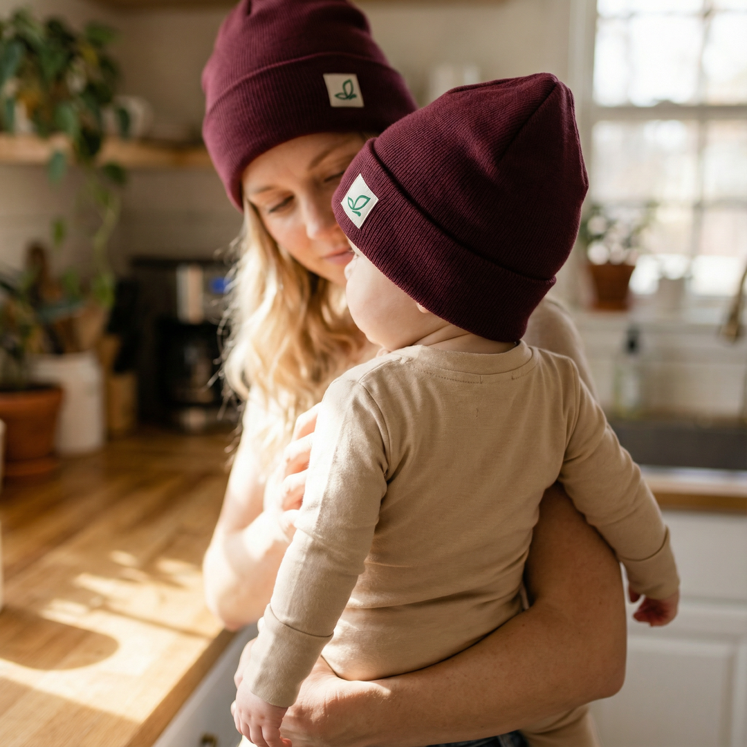 Mutter hält Baby in heller Küche, beide tragen bordeauxrote Bio-Babymützen von Elsara